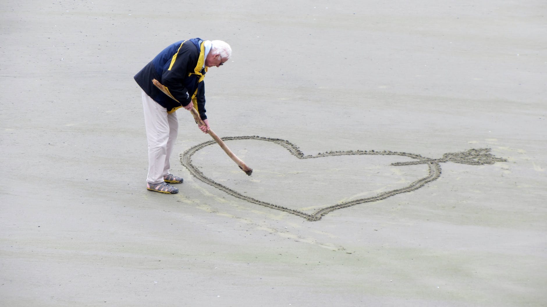 man wearing blue jacket holding a brown stick towards the heart drawn on sand